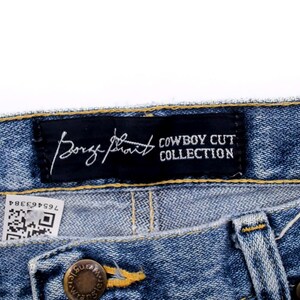 Pu&ograve; includere: Primo piano di jeans blu. Un'etichetta nera recita "George Strait COWBOY CUT COLLECTION" in testo bianco. I jeans hanno una chiusura con bottone e cerniera. &Egrave; visibile una piccola etichetta con un codice QR.