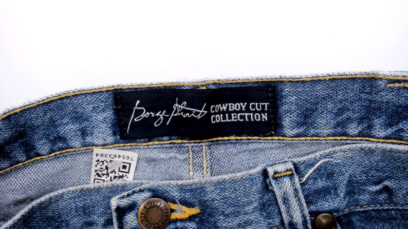 Pu&ograve; includere: Primo piano di jeans blu. Un'etichetta nera recita "George Strait COWBOY CUT COLLECTION" in testo bianco. I jeans hanno una chiusura con bottone e cerniera. &Egrave; visibile una piccola etichetta con un codice QR.