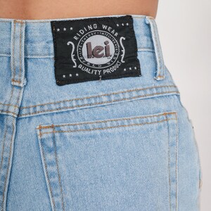Puede incluir: Vaqueros de mezclilla azul claro con un parche negro en el bolsillo trasero que dice "Riding Wear - Life Energy - Intelligence - World Quality Product - Lei".