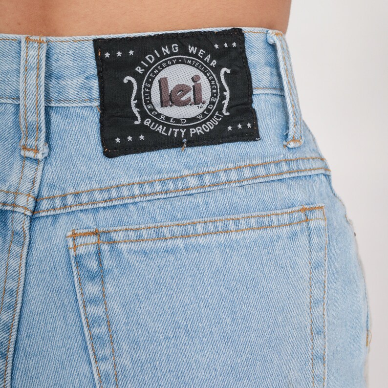 Puede incluir: Vaqueros de mezclilla azul claro con un parche negro en el bolsillo trasero que dice "Riding Wear - Life Energy - Intelligence - World Quality Product - Lei".