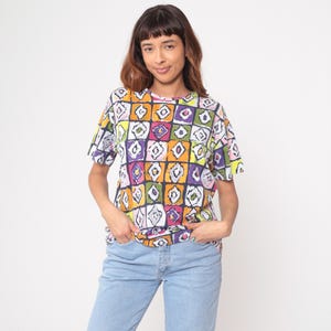 May include: A short-sleeved, colorful t-shirt with a geometric pattern of squares in yellow, orange, purple, green, and white. The shirt is paired with light blue jeans. The model is smiling and has their hands in their pockets.