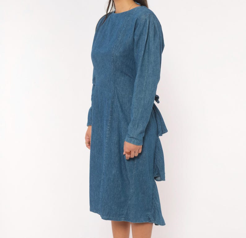 Puede incluir: Un vestido de mezclilla de manga larga en un tono azul medio. El vestido presenta un cuello redondo, un corpi&ntilde;o ajustado con detalles de costuras verticales y una falda que llega hasta la rodilla. La tela tiene un tono azul uniforme.