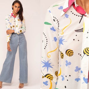 May include: A white button-down shirt with a colorful pattern of bees, flowers, and abstract shapes. The collar and cuffs have a pink and red trim. The shirt is paired with light blue wide-leg jeans.