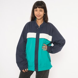 Könnte beinhalten: Eine marineblaue, türkisfarbene und weiße Windjacke. Die Jacke hat einen Stehkragen und einen durchgehenden Reißverschluss. Die Jacke hat ein Colorblock-Design mit einem weißen Streifen über der Brust und türkisfarbenen Einsätzen auf der unteren Hälfte.