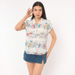 May include: A short-sleeved button-down shirt in white featuring a colorful hand-drawn style print of various buildings and houses in shades of red, blue, yellow, and green, paired with a dark blue denim mini skirt with a side slit.