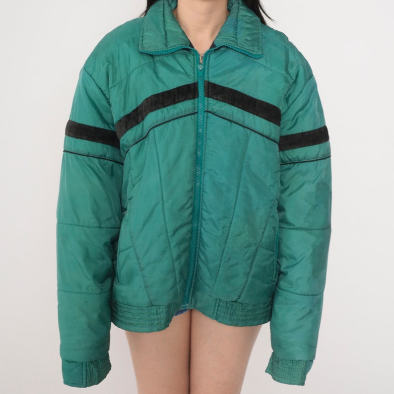 May include: A teal blue puffer jacket with black and brown stripes on the shoulders and sleeves. The jacket has a zipper closure and a stand-up collar.