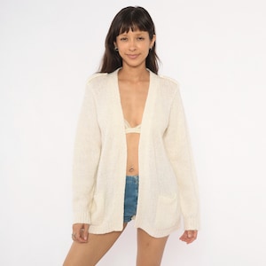 May include: A long-sleeved, open-front cardigan in a solid cream color. This knit garment features a ribbed texture along the front edges, cuffs, and hem, along with two patch pockets positioned on the lower front section of the piece.