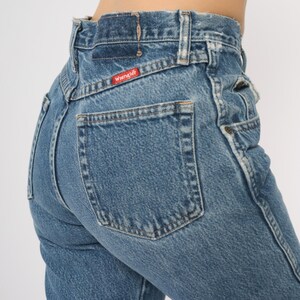 Puede incluir: Primer plano de unos vaqueros Wrangler de mezclilla azul. Los vaqueros tienen cintura alta, un bolsillo trasero y el logotipo de Wrangler en una etiqueta roja. La mezclilla est&aacute; descolorida y las costuras son visibles.
