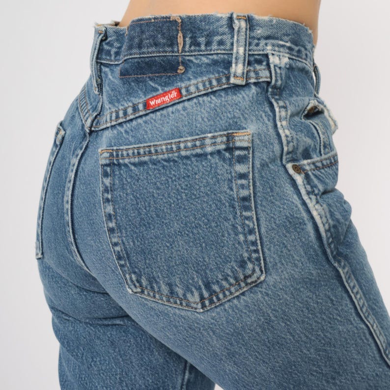 Puede incluir: Primer plano de unos vaqueros Wrangler de mezclilla azul. Los vaqueros tienen cintura alta, un bolsillo trasero y el logotipo de Wrangler en una etiqueta roja. La mezclilla est&aacute; descolorida y las costuras son visibles.