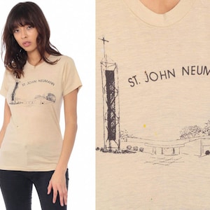 May include: Tan short-sleeve t-shirt featuring a graphic of a church with a tall steeple and the text "ST. JOHN NEUMANN" in black. The shirt is worn by a person wearing black pants.