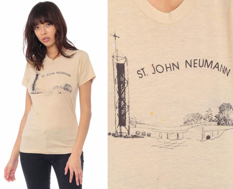 May include: Tan short-sleeve t-shirt featuring a graphic of a church with a tall steeple and the text "ST. JOHN NEUMANN" in black. The shirt is worn by a person wearing black pants.