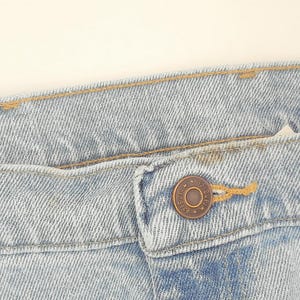 May include: Close-up of light blue denim jeans. The jeans have a button closure and a metal button. The stitching is visible, and the fabric has a textured appearance. A hand with red nail polish is holding the jeans.