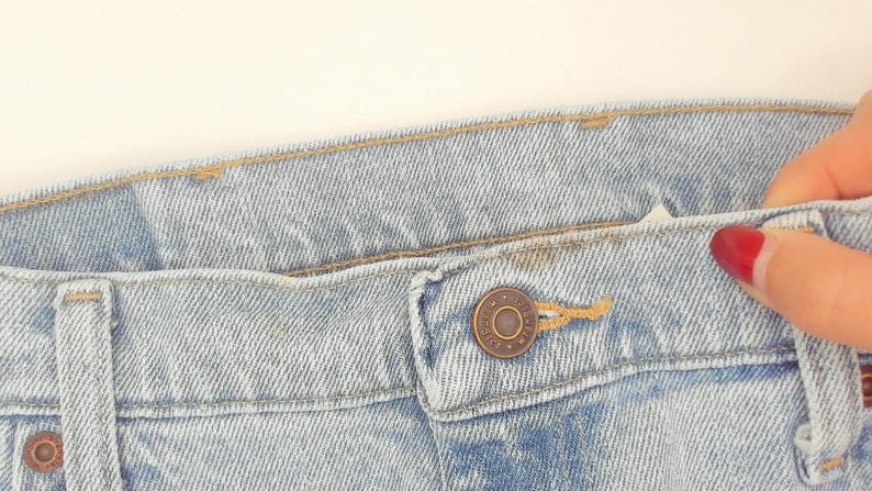 May include: Close-up of light blue denim jeans. The jeans have a button closure and a metal button. The stitching is visible, and the fabric has a textured appearance. A hand with red nail polish is holding the jeans.