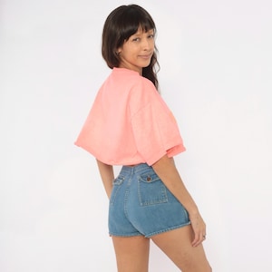 K&ouml;nnte beinhalten: Ein leuchtend korallenrosa kurz&auml;rmeliges bauchfreies Oberteil kombiniert mit einer hoch taillierten blauen Jeansshorts. Die Shorts verf&uuml;gen &uuml;ber zwei markante aufgesetzte Taschen auf der R&uuml;ckseite, die jeweils mit einem Knopf verschlossen sind.