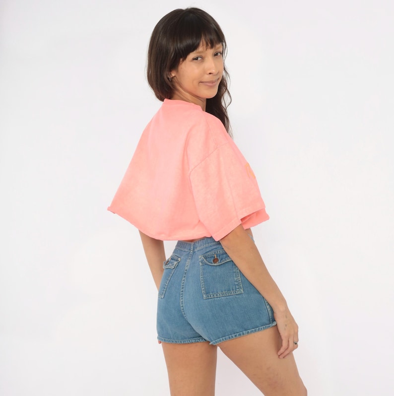 K&ouml;nnte beinhalten: Ein leuchtend korallenrosa kurz&auml;rmeliges bauchfreies Oberteil kombiniert mit einer hoch taillierten blauen Jeansshorts. Die Shorts verf&uuml;gen &uuml;ber zwei markante aufgesetzte Taschen auf der R&uuml;ckseite, die jeweils mit einem Knopf verschlossen sind.