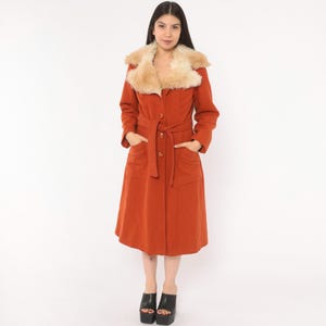 May include: A long, burnt orange coat featuring a large, fluffy cream-colored faux fur collar. The coat has long sleeves, a matching fabric tie belt at the waist, and two large patch pockets on the front. It is fastened with three visible buttons.