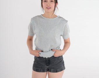 May include: A woman wearing a silver sparkly short-sleeved shirt and black denim shorts.