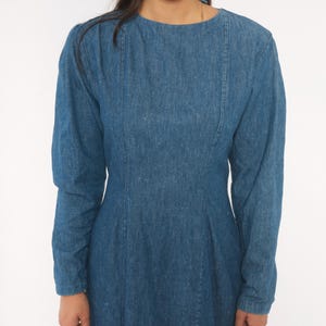 Puede incluir: Un vestido de mezclilla de manga larga en un tono azul medio, con cuello redondo, costuras verticales que van desde los hombros hasta la cintura y un corpi&ntilde;o ajustado que se transforma en una falda ligeramente acampanada.