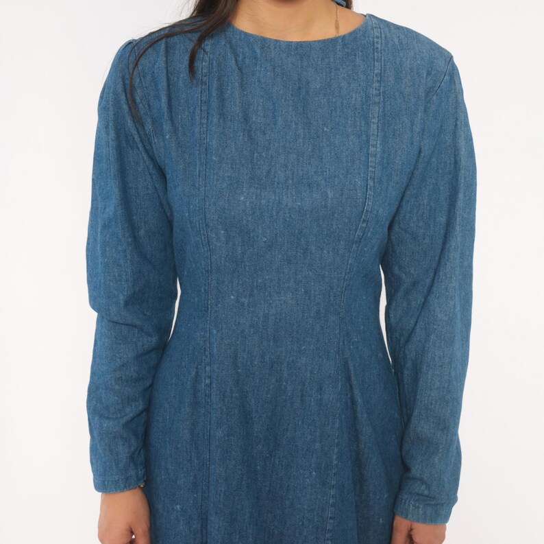 Puede incluir: Un vestido de mezclilla de manga larga en un tono azul medio, con cuello redondo, costuras verticales que van desde los hombros hasta la cintura y un corpi&ntilde;o ajustado que se transforma en una falda ligeramente acampanada.