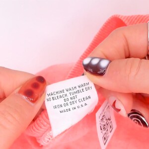 K&ouml;nnte beinhalten: Ein wei&szlig;es, rechteckiges Pflegeetikett an einem korallenrosa Stoff. Der Text lautet: MACHINE WASH WARM, NO BLEACH, TUMBLE DRY, DO NOT IRON OR DRY CLEAN, MADE IN U.S.A. Ein zweites, kleineres wei&szlig;es Etikett mit einem QR-Code ist teilweise sichtbar.