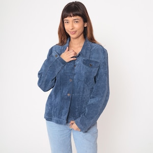 May include: A blue suede jacket with button closures and two chest pockets. The jacket is paired with light blue jeans. The jacket has a classic, casual style.