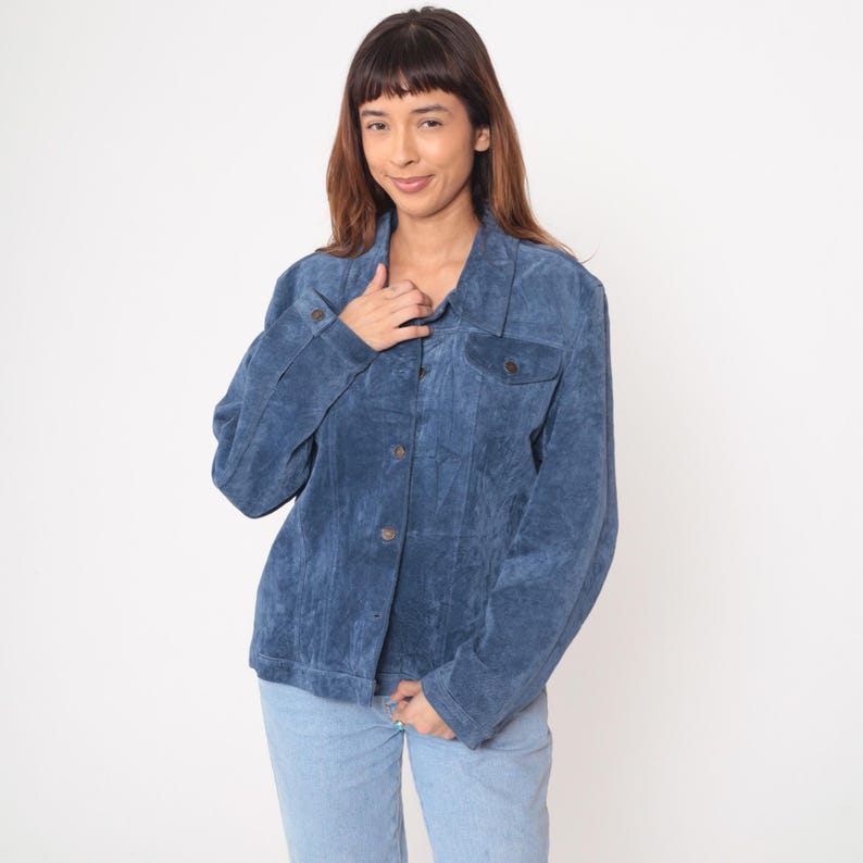 May include: A blue suede jacket with button closures and two chest pockets. The jacket is paired with light blue jeans. The jacket has a classic, casual style.