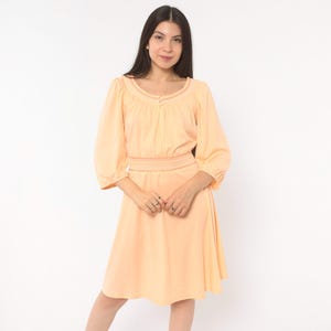 May include: A peach-colored knee-length dress featuring three-quarter length puff sleeves, a gathered scoop neckline with a small central button detail, and a wide smocked elasticized waistband that creates a defined silhouette.