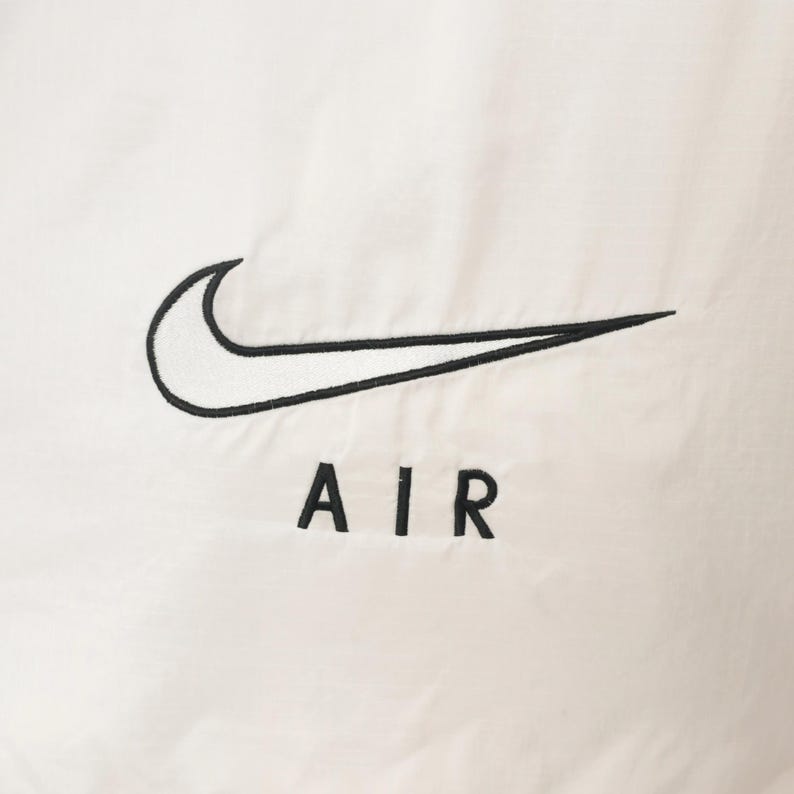 Puede incluir: Primer plano de una tela blanca con el logotipo de Nike swoosh bordado en negro y la palabra "AIR" debajo. El logotipo es una marca de verificaci&oacute;n curva y el texto est&aacute; en una fuente sans-serif.
