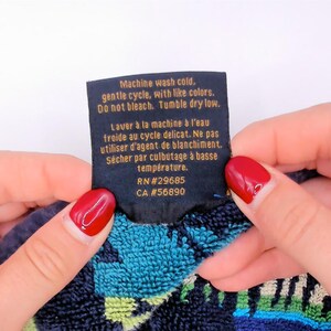 May include: A black fabric care label with gold text reading: Machine wash cold, gentle cycle, with like colors. Do not bleach. Tumble dry low. RN#29685 CA #56890. The label is attached to a textured fabric with blue, green, and black patterns.