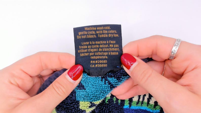 May include: A black fabric care label with gold text reading: Machine wash cold, gentle cycle, with like colors. Do not bleach. Tumble dry low. RN#29685 CA #56890. The label is attached to a textured fabric with blue, green, and black patterns.