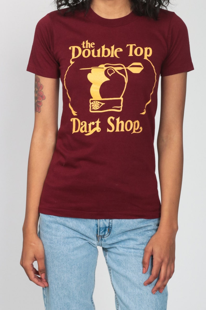 May include: A maroon t-shirt with a gold graphic of a hand throwing a dart at a dartboard. The graphic says "the Double Top Dart Shop."
