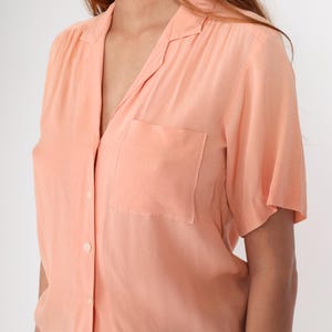 Puede incluir: Una camisa abotonada de color melocot&oacute;n con mangas cortas y un bolsillo en el pecho. La camisa tiene un cuello cl&aacute;sico y un corte relajado, adecuado para ropa informal. La tela parece ligera y ligeramente texturizada.