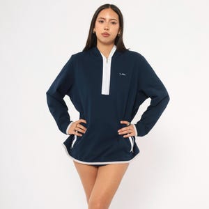 May include: Navy blue pullover with a white zipper and trim. The long-sleeved top has a drawstring at the waist and a small logo on the chest. The collar and cuffs are white. The model is wearing the top.