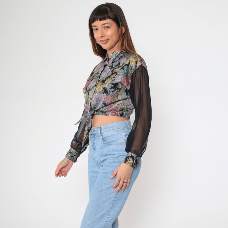 May include: A floral print blouse with sheer black sleeves and a tied front. The blouse features a button-down design and is paired with light blue jeans. The floral pattern includes shades of pink, purple, and green on a black background.