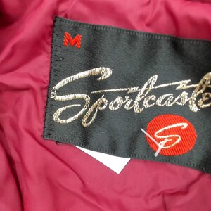 May include: Close-up of a black label with the embroidered word "Sportcaster" in gold script. A red circle with a white "S" is below the script. The label is sewn onto a burgundy fabric, with a red "M" at the top.