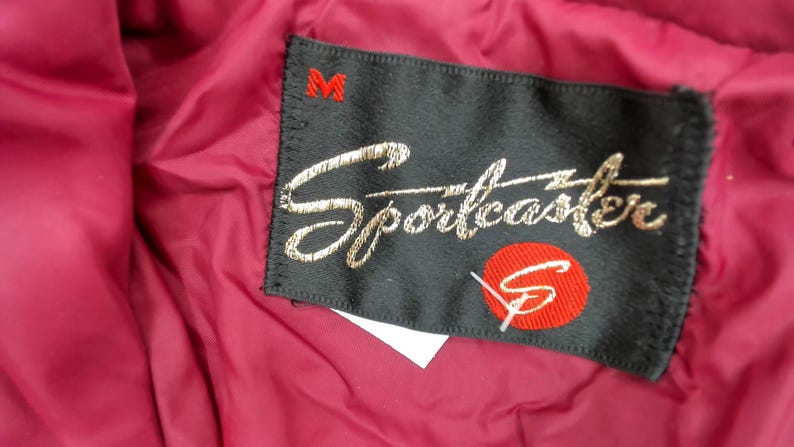 May include: Close-up of a black label with the embroidered word "Sportcaster" in gold script. A red circle with a white "S" is below the script. The label is sewn onto a burgundy fabric, with a red "M" at the top.