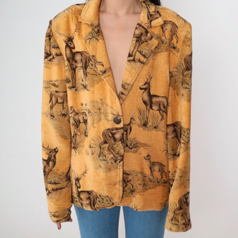 May include: Mustard-colored blazer with a deer pattern. The blazer features a single button closure and a classic lapel design. The deer are depicted in a brown and black color scheme, set against a landscape background.