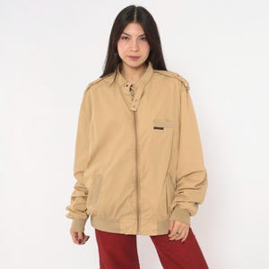 May include: A tan zip-up jacket featuring a high collar with a snap button closure, shoulder epaulets, long sleeves with ribbed cuffs, and a small chest pocket. The jacket has a ribbed hem and a central zipper closure, providing a structured fit.