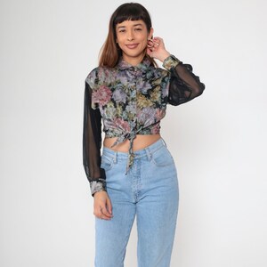 May include: A floral print blouse with sheer black sleeves and a tie-front detail. The blouse features a button-down collar and a mix of pink, purple, and green floral patterns on a black background. Paired with light blue jeans.