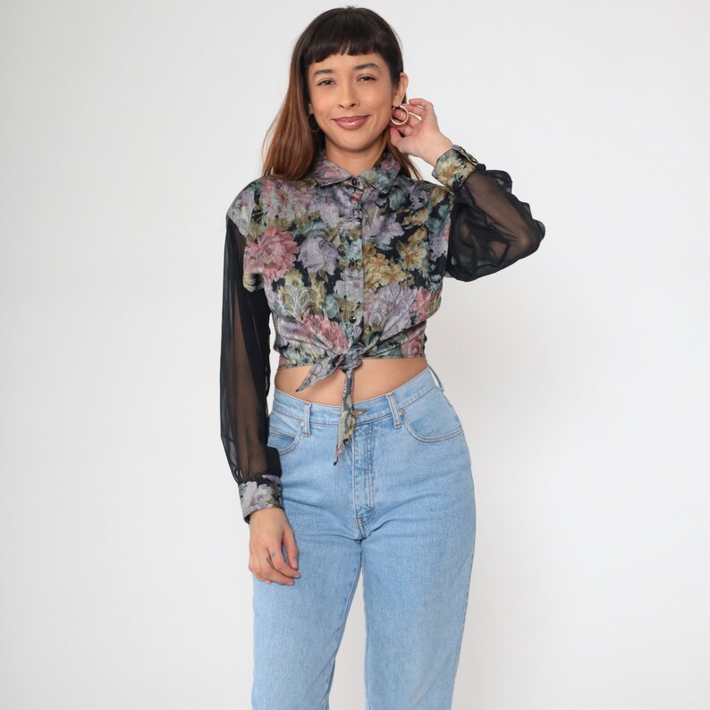 May include: A floral print blouse with sheer black sleeves and a tie-front detail. The blouse features a button-down collar and a mix of pink, purple, and green floral patterns on a black background. Paired with light blue jeans.