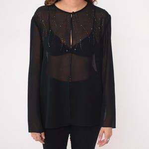 May include: Black sheer long-sleeve blouse with a round neckline and a keyhole opening. The blouse features delicate beaded embellishments along the neckline and upper chest. The garment is paired with black pants.