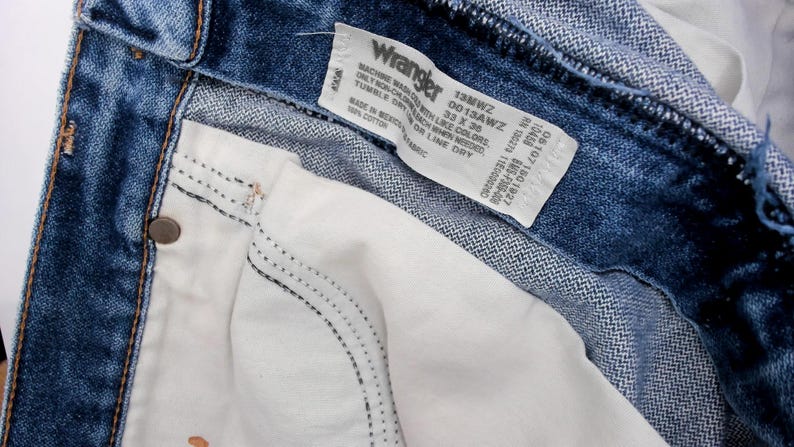 May include: Close-up of blue denim Wrangler jeans. The jeans have a white interior pocket and a label with the Wrangler logo and size information. The jeans are size 33 x 30.