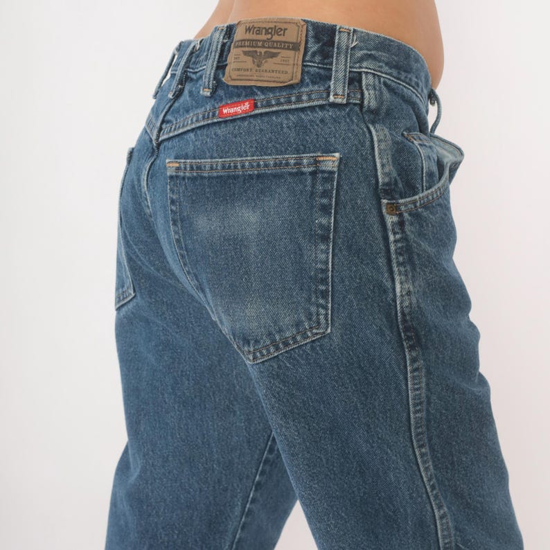 May include: Blue denim Wrangler jeans. The jeans have a classic five-pocket design, with a small red Wrangler tag on the back pocket. The Wrangler logo is visible on the leather patch at the waistband. The jeans are a medium wash.