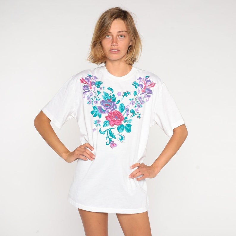 May include: White t-shirt with a floral design in shades of pink, purple, and teal.