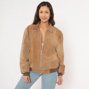May include: A tan suede jacket with a zip-up front, collar, and brown ribbed cuffs and hem. The jacket is worn over a white top and paired with blue jeans. The model is smiling.