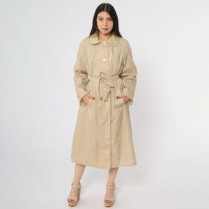 May include: A beige trench coat featuring a button closure, a tie belt, and pockets. The coat has a classic collar and long sleeves. The model is wearing platform sandals. The coat is knee-length and made of a lightweight material.