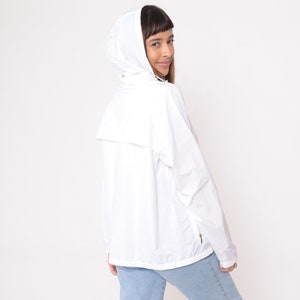 May include: A white hooded jacket with a zipper closure and a small pocket on the front. The jacket is made of a lightweight material and has a relaxed fit. The model is wearing light blue jeans.