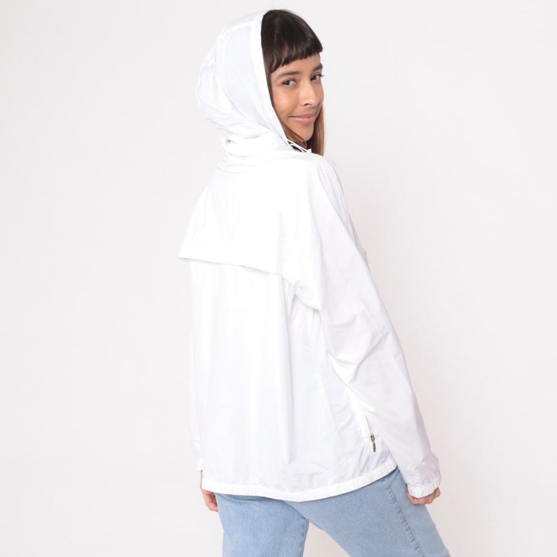 May include: A white hooded jacket with a zipper closure and a small pocket on the front. The jacket is made of a lightweight material and has a relaxed fit. The model is wearing light blue jeans.