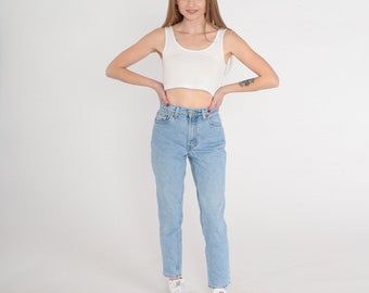 May include: A woman wearing a white tank top and light blue denim jeans. She is standing with her hands on her hips and is wearing white sneakers.