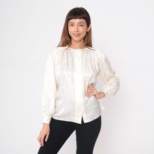 May include: Cream-colored long-sleeve blouse with a subtle, swirling pattern. The blouse features a button-up front and a rounded neckline. The sleeves have a slight puff at the shoulders and gather at the wrists. The model is wearing black pants.